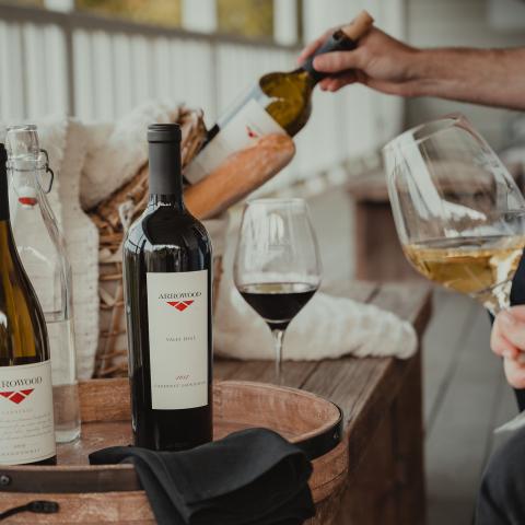 A basket of bread with two bottles of Arrowood wines on a tray, and two people holding a glass of red and white wine.