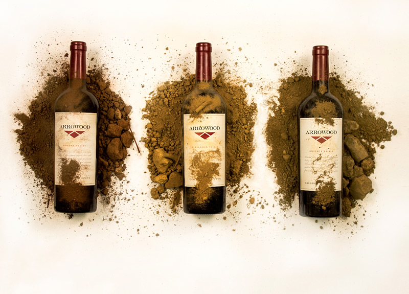 Three bottles of Arrowood Vineyard's wine laying down flat, each in a dirt pile of their unique soil type.